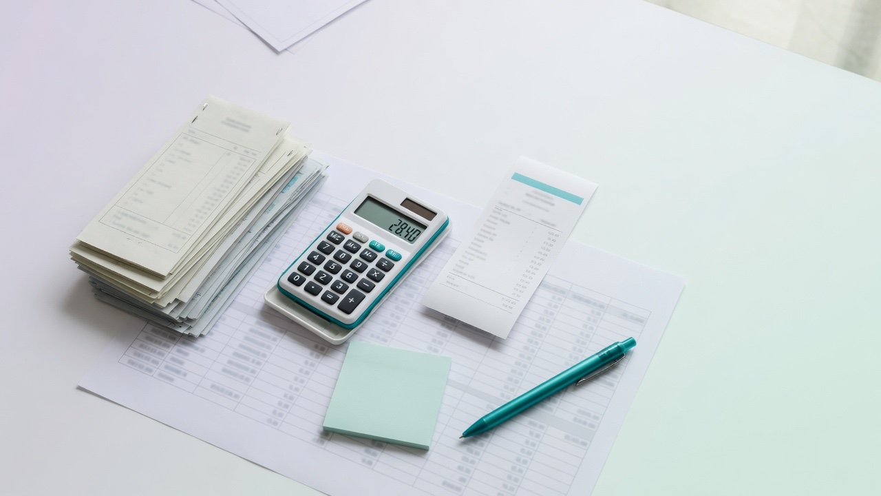 Top-down light desk: tall stack of subscription invoices, one thin pay-per-use receipt, pocket calculator, mint sticky note, and teal pen on a comparison sheet